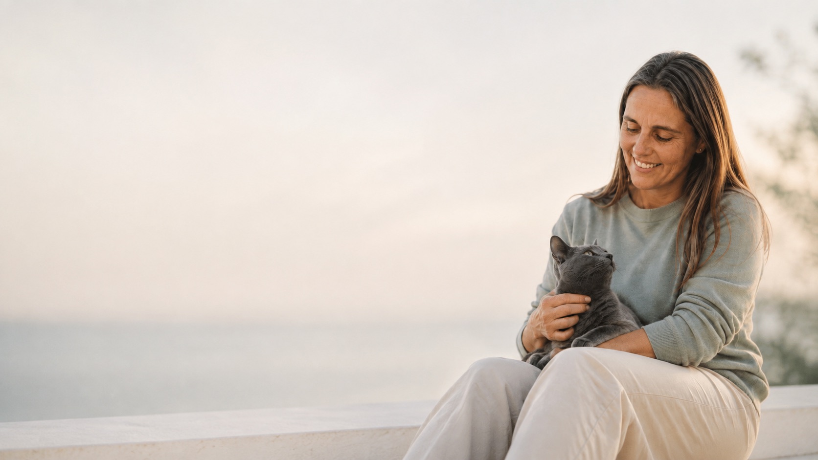 Eva Español sentada al atardecer junto al mar, con un gato azul en el regazo